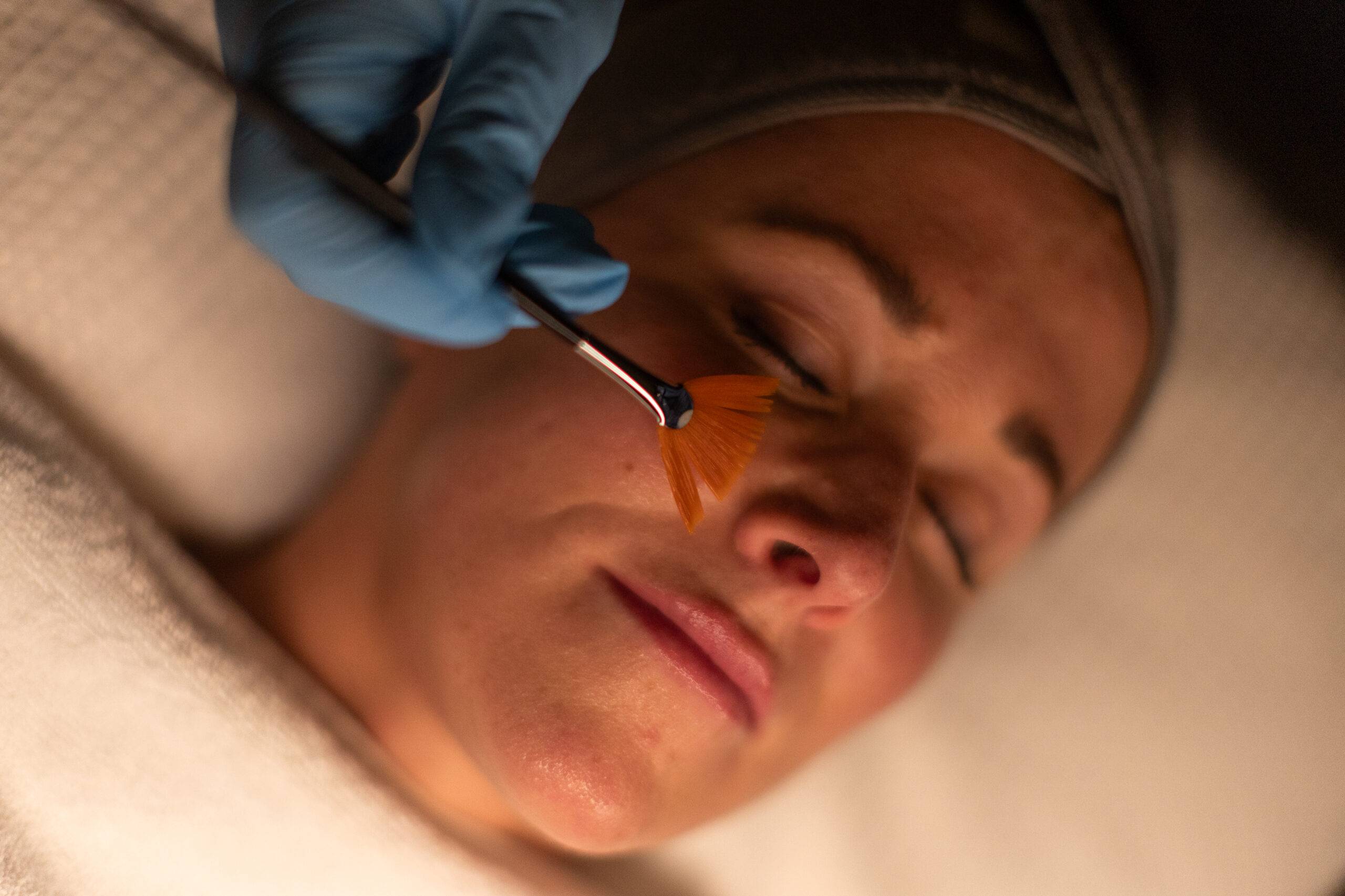 A woman receiving a medifacial treatment, with her eyes closed and a brush applying product to her face. The aesthetician wears blue gloves. The setting is warmly lit, focusing closely on the treatment