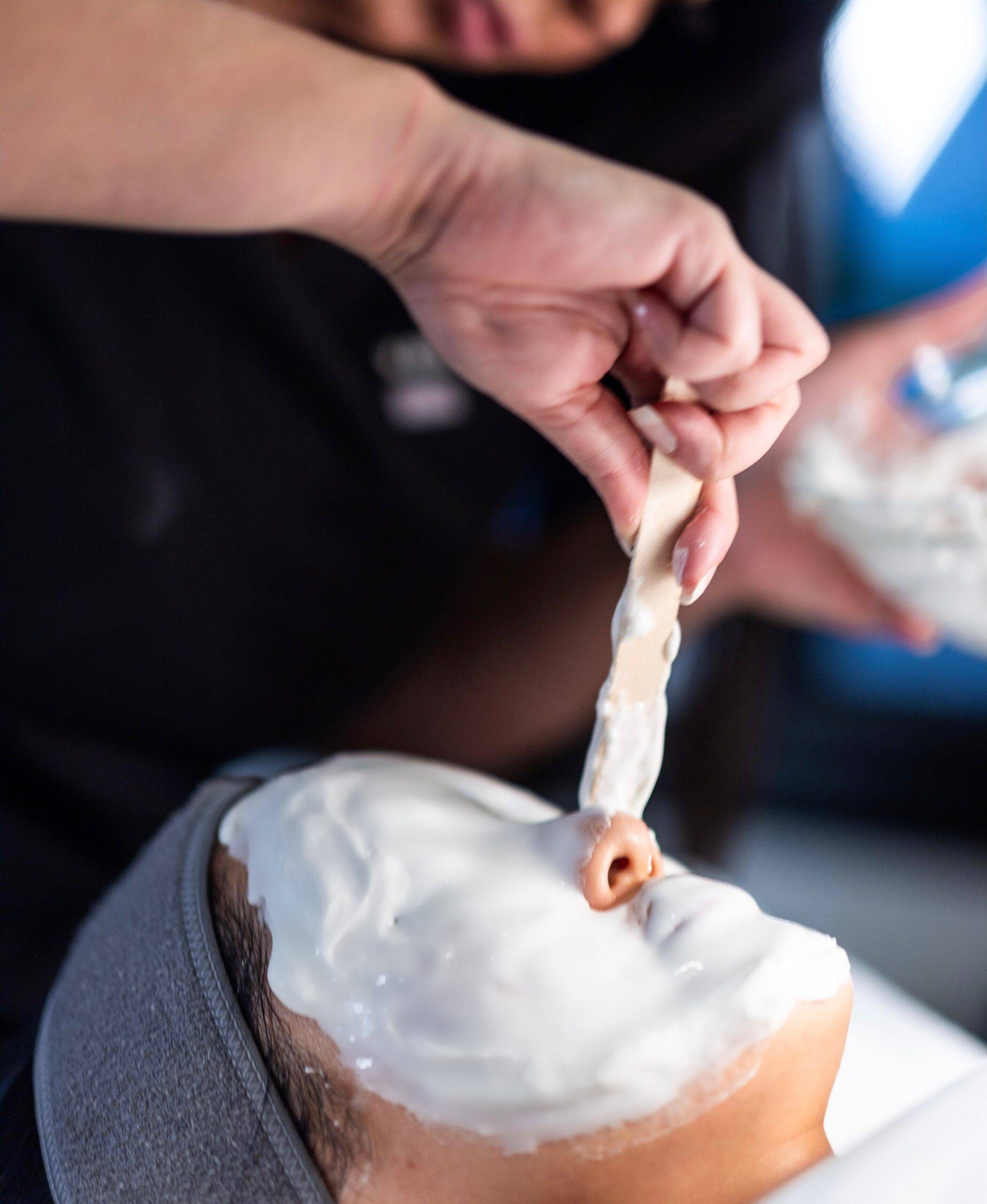 A person applies a thick layer of white cream on their face using a spatula, with focus on their hands and the skin's surface, as part of a facial rejuvenation medifacial treatment.