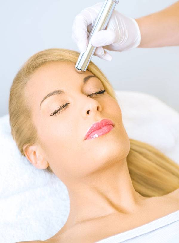 A woman receiving a microdermabrasion treatment with a modern skin-care device, lying calmly with eyes closed. The aesthetician, wearing gloves, uses the device on her forehead in a clinical setting
