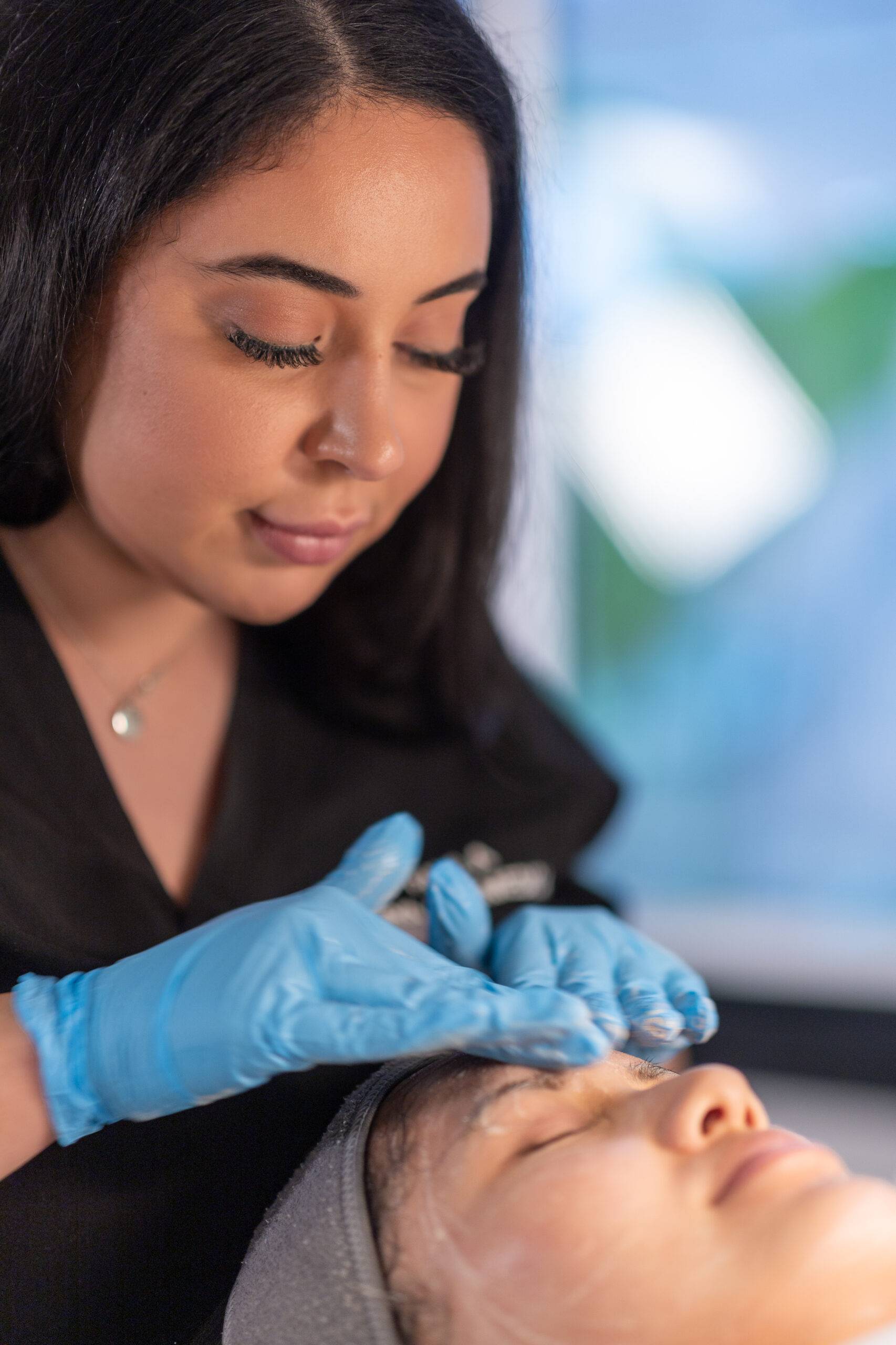 A beautician in black attire and blue gloves performs a skin treatment on a relaxed female client lying down, focusing intently on her task in a brightly lit salon setting.