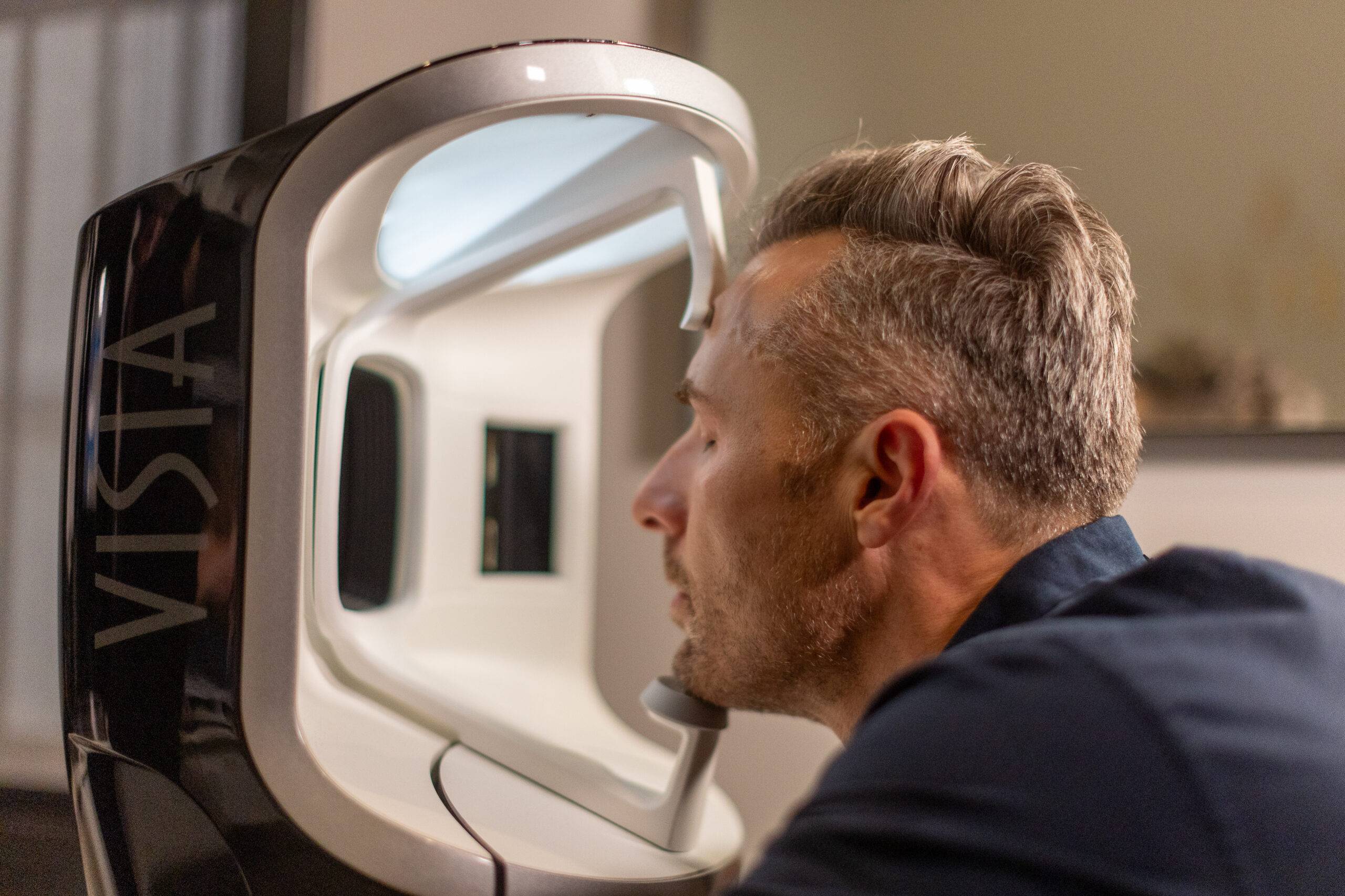 A man with blond hair closely examines the screen of a large, modern, rectangular Visia Skin Analysis machine labeled "vista" in a dimly lit room.