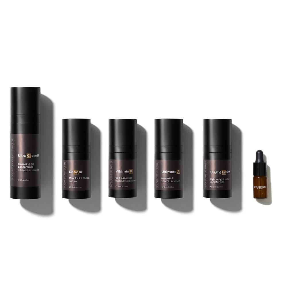 Six skincare products from the Synergie Balance + Clarity Kit in black containers are arranged in a line on a light gray background, varying in size and labeled with their respective functions, such as