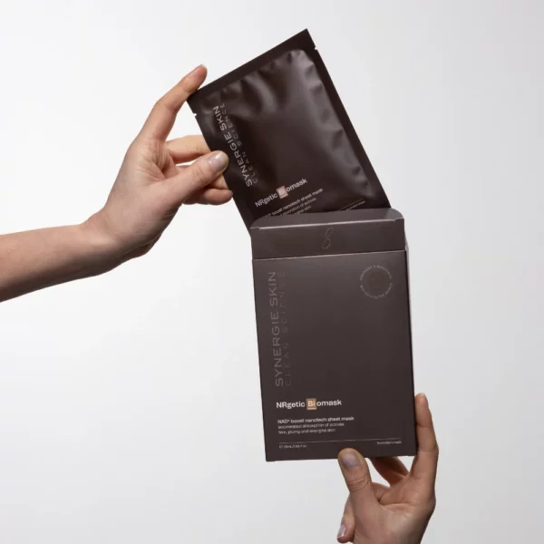 A person holds a box of NRgetic Biomask by Synergie Skin in one hand, while using the other to take out a packet. The packaging is elegantly dark and minimalist, highlighted by bold silver lettering. A plain white background enhances the product's sleek design.