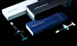 Three boxes labeled "Rejuran" in white, black, and blue rest on a sleek black surface. Nearby, two syringes—with a teal and a black plunger—hint at advanced skin regeneration solutions. The setup exudes an air of innovative medical or cosmetic excellence.